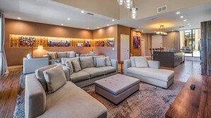 A modern living room with a sectional sofa, ottoman, and a wooden coffee table. There is a dining area with a table and chairs, and a kitchen in the background. - Mvv7482 - 3BR House Near Disney at Magic Village (Kissimmee)