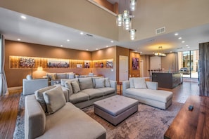 Ein modernes Wohnzimmer mit Ecksofa, Hocker und Couchtisch. Es gibt einen Essbereich mit Tisch und Stühlen, und im Hintergrund eine Küche.