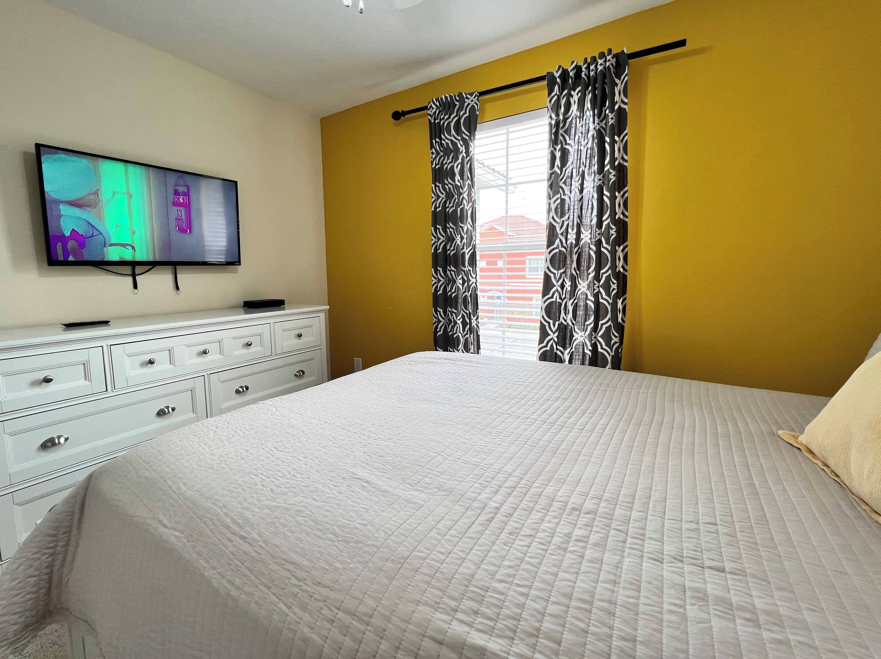 Une chambre à coucher avec un lit, une commode, un téléviseur fixé au mur et des rideaux à motifs.