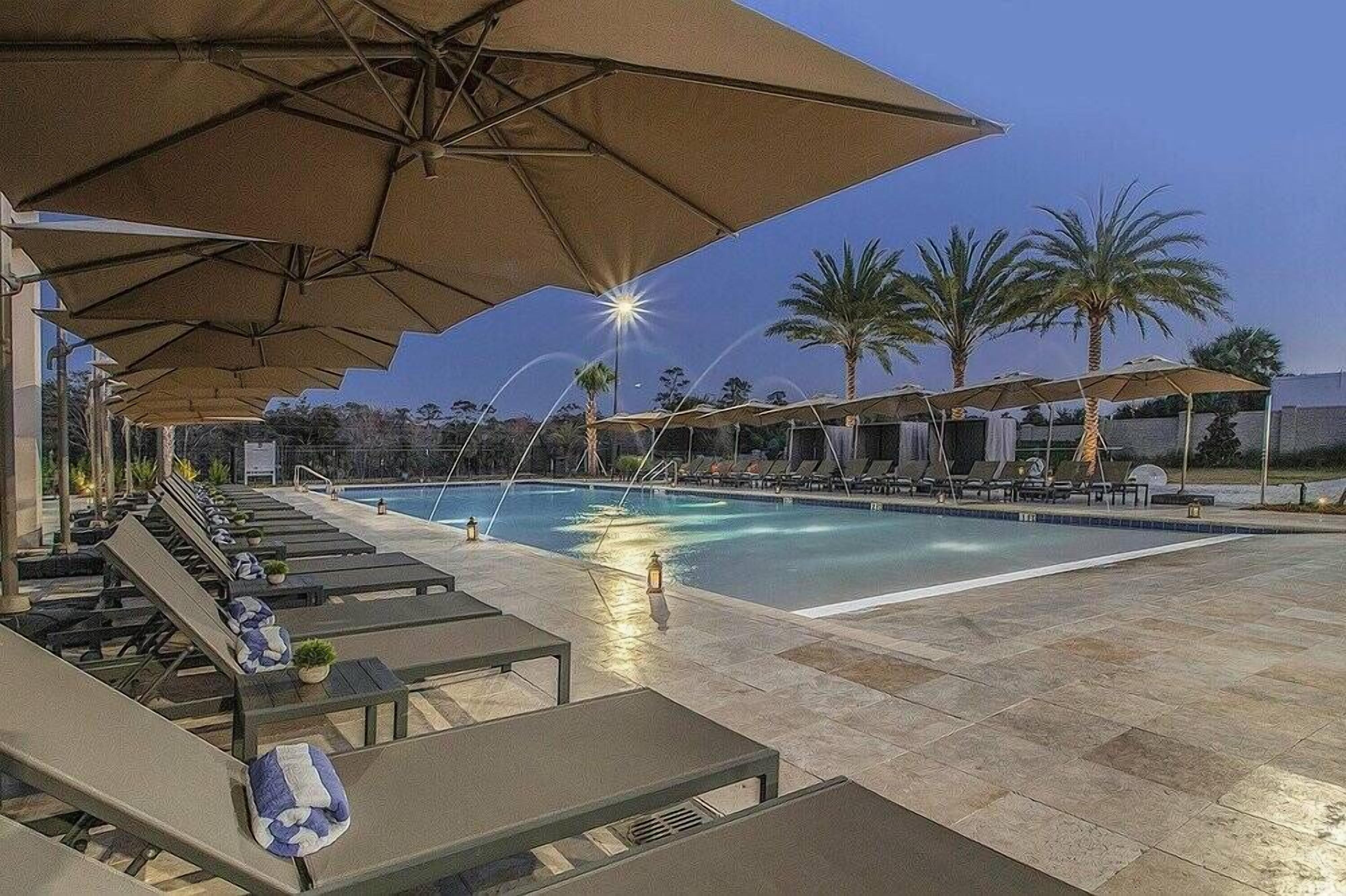A poolside area with lounge chairs and umbrellas, a pool, and palm trees in the background.