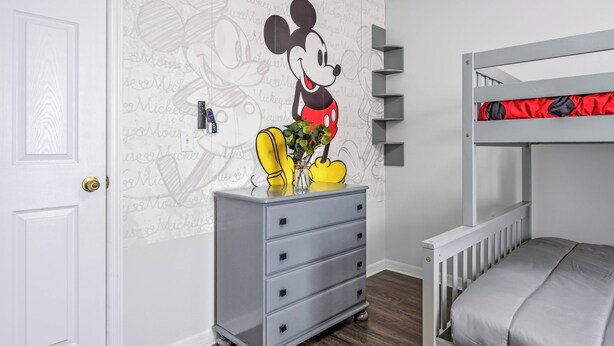 Une chambre d’enfant avec un lit superposé, une commode grise et un papier peint à l’effigie de Mickey Mouse.