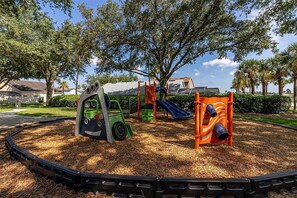 Une aire de jeux avec un toboggan vert et orange, une structure d’escalade et un espace de jeux sur le thème du train.
