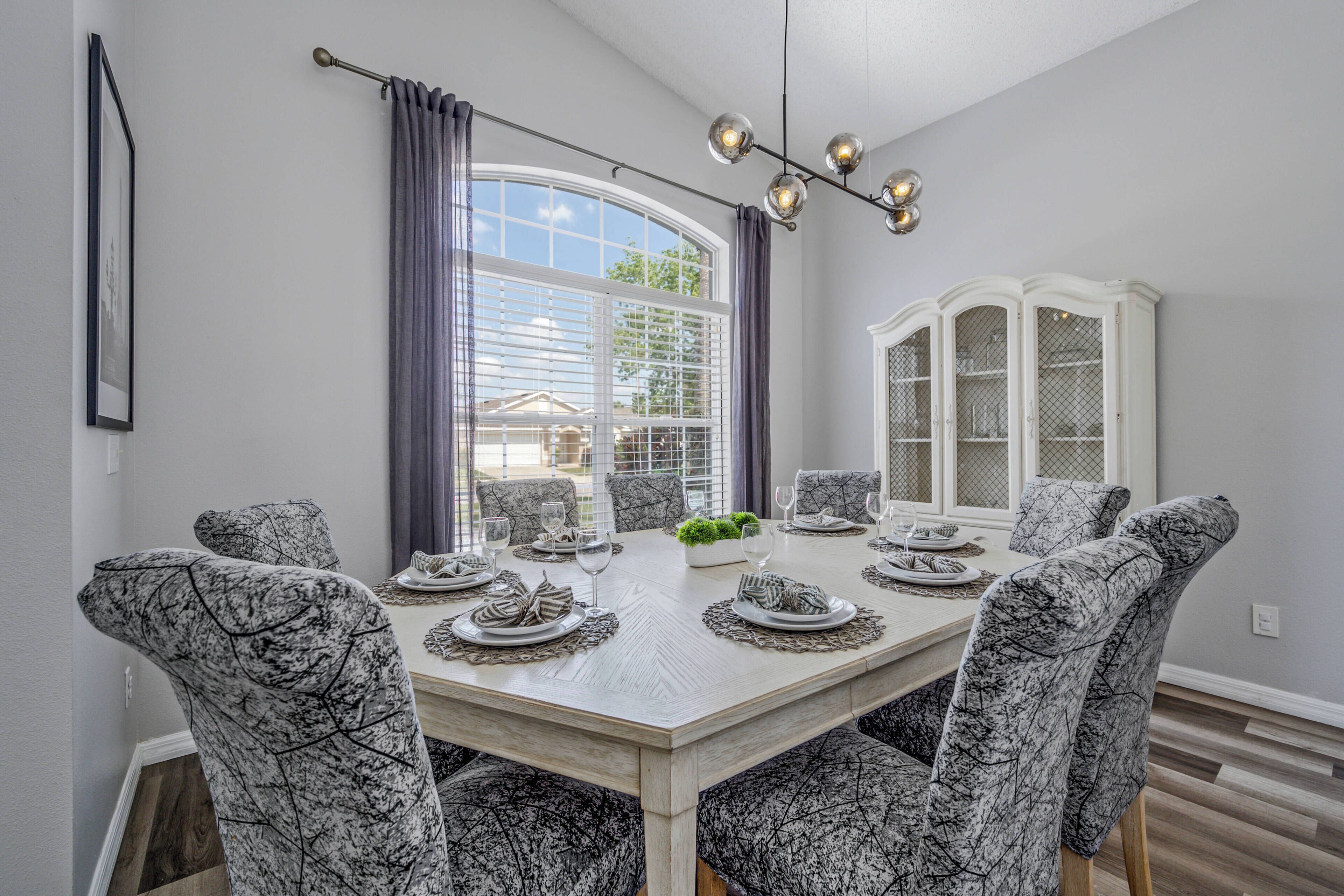 A dining room with a large table set for eight, surrounded by patterned chairs, a white cabinet, and a window with curtains.