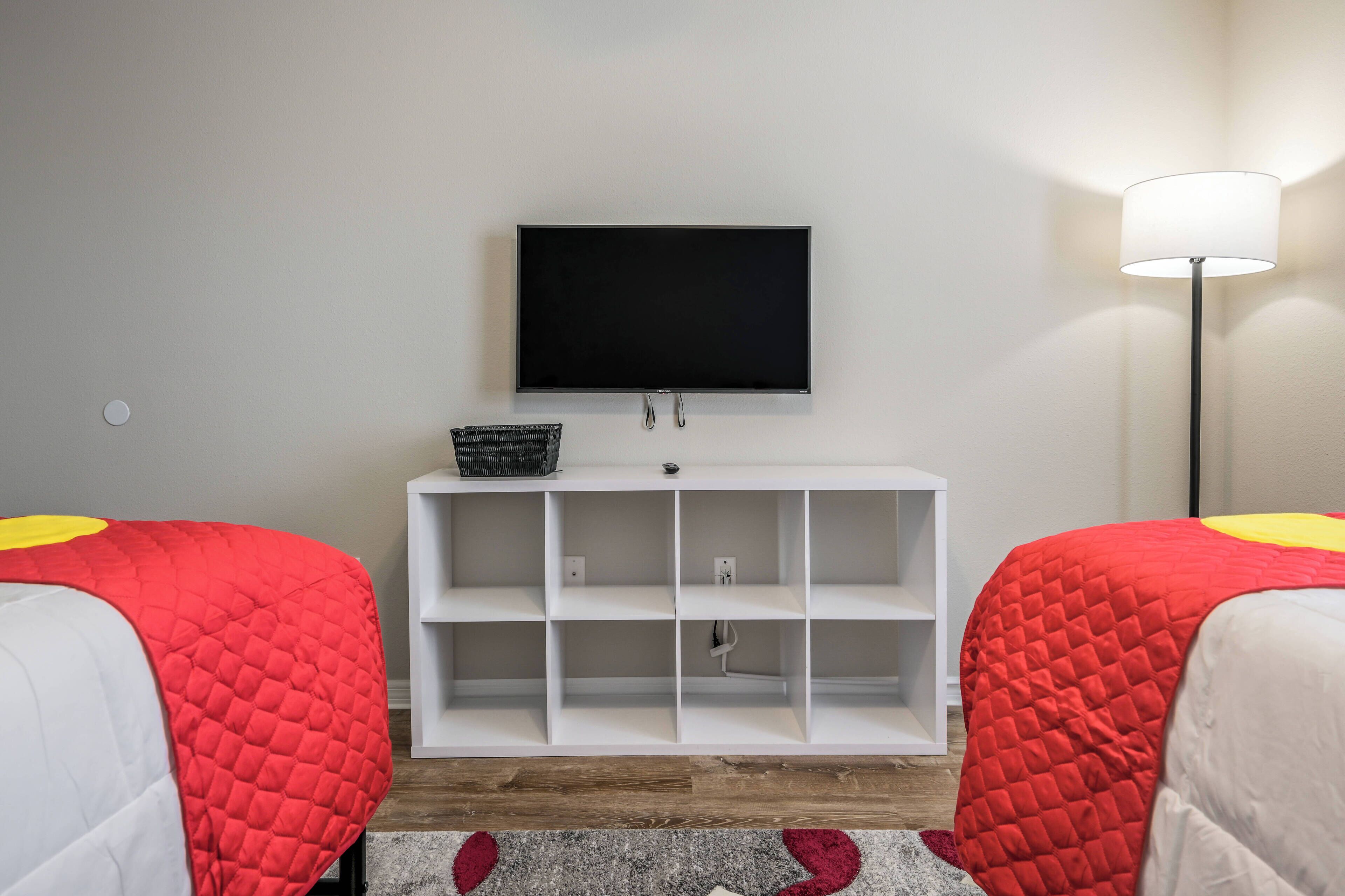 A room with two beds, a TV mounted on the wall, a white shelving unit, and a floor lamp.