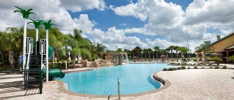 A swimming pool with a water slide, palm trees, and a brick patio.