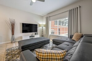 A modern living room with a sectional sofa, a flat-screen TV, a lamp, and a view of a building through a window. - Pp8968 - 4BR House With Pool Near Disney Parks (Kissimmee)