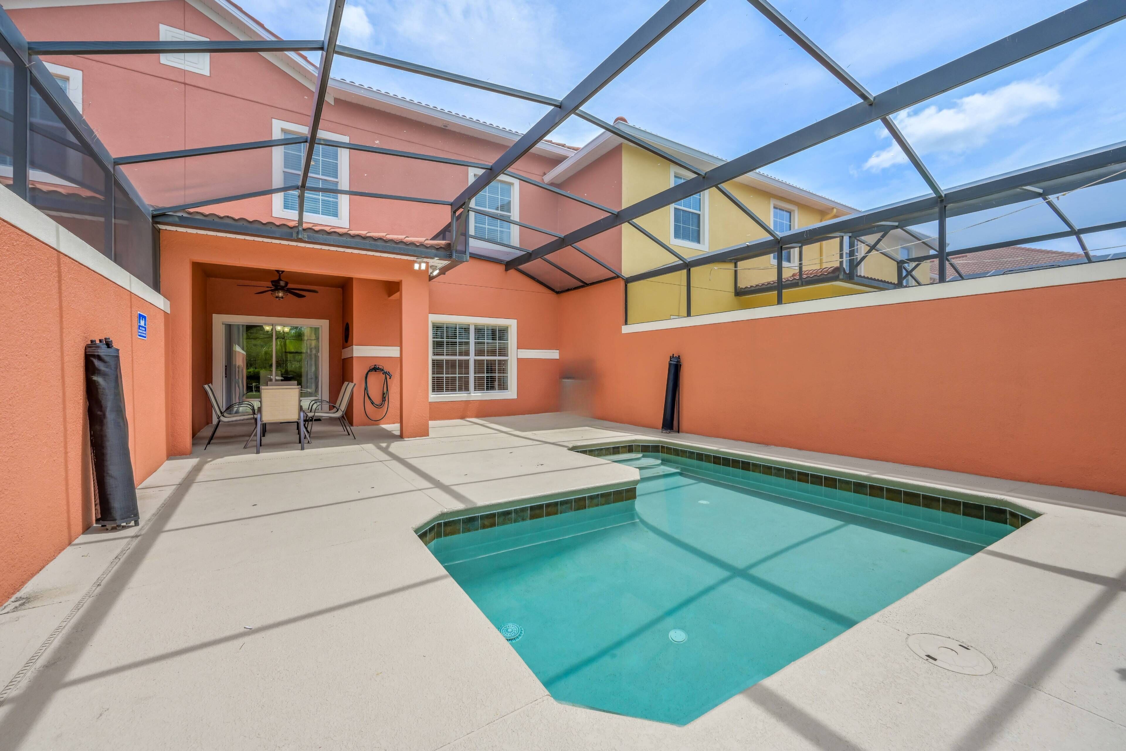 A covered swimming pool area with a ceiling structure, a door leading to an outdoor space, and a table with chairs.