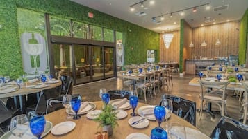 A restaurant with round tables set with blue glassware, white plates, and silverware. There are chairs around the tables, and a potted plant on one table. The walls are green with a leafy pattern, and there is a large window in the background.