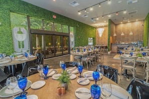 A restaurant with round tables set with blue glassware, white plates, and silverware. There are chairs around the tables, and a potted plant on one table. The walls are green with a leafy pattern, and there is a large window in the background. - Mvv7460 - 3BR House Near Disney w/ Hot tub (Kissimmee)