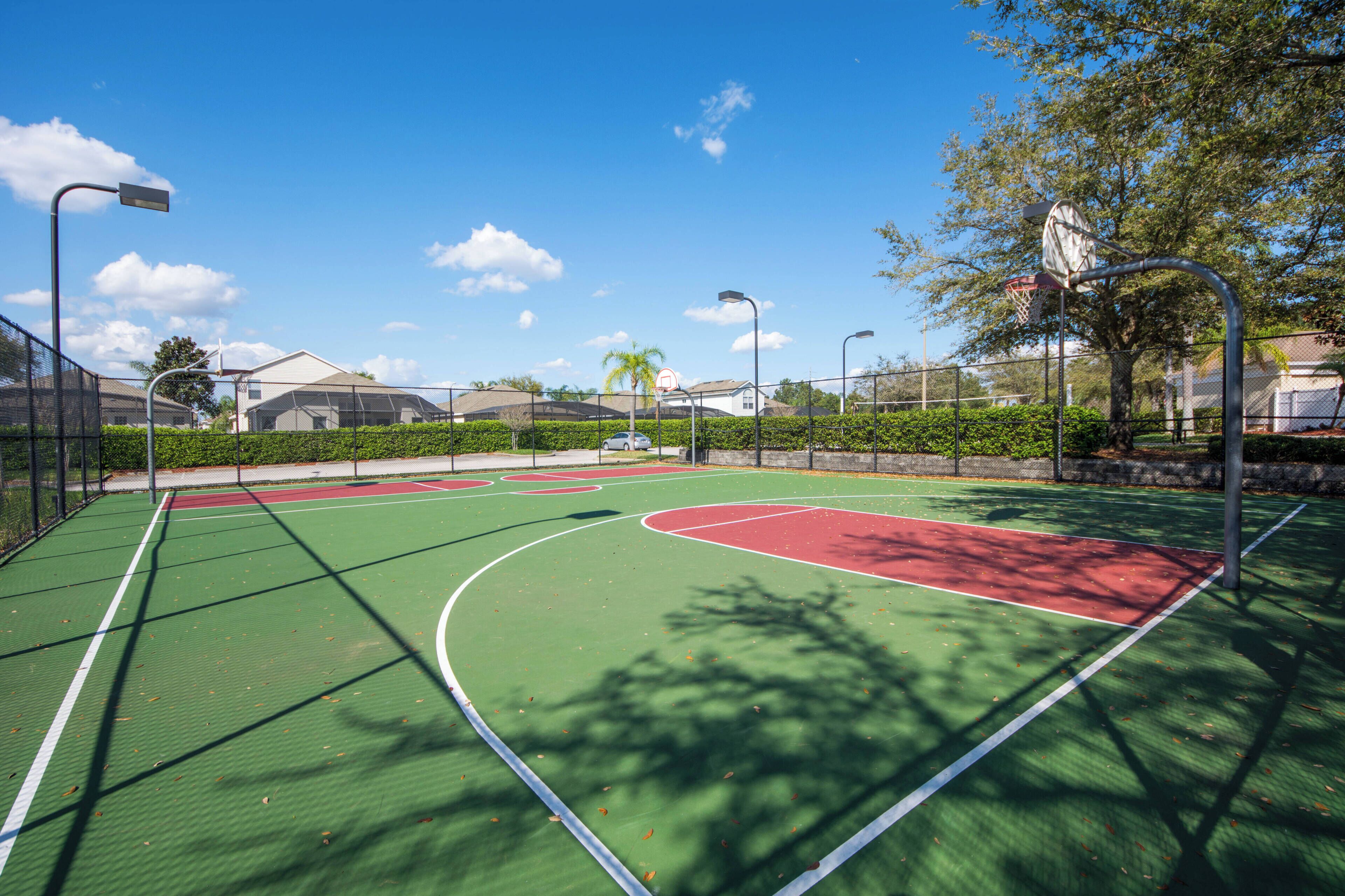 An outdoor basketball court with green and red surfaces, surrounded by a fence, street lamps, and trees.