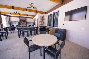 Un área de comedor al aire libre con mesas y sillas redondas, un ventilador de techo y un televisor montado en la pared.