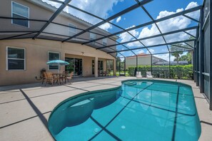 Piscina exterior con cerramiento de vidrio, zona de estar cubierta con sillas y vista a casas vecinas.