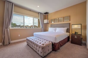 Ein Schlafzimmer mit einem großen Bett, einem Fußhocker mit Knopfauflage, einem Nachttisch mit Lampe und einem Fenster mit Vorhängen.