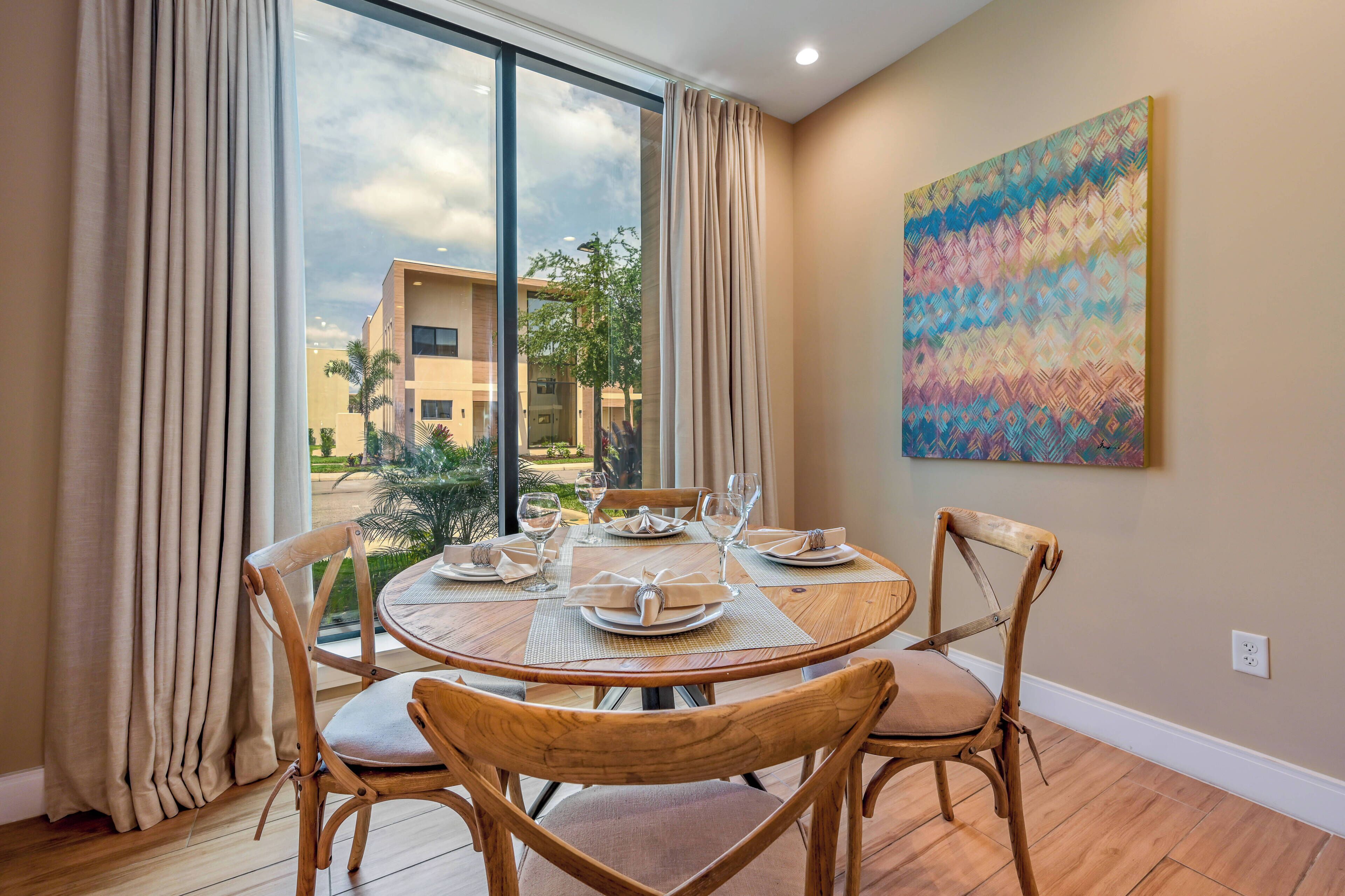 A dining area with a round table set for four, wooden chairs, and a view of buildings and greenery outside.