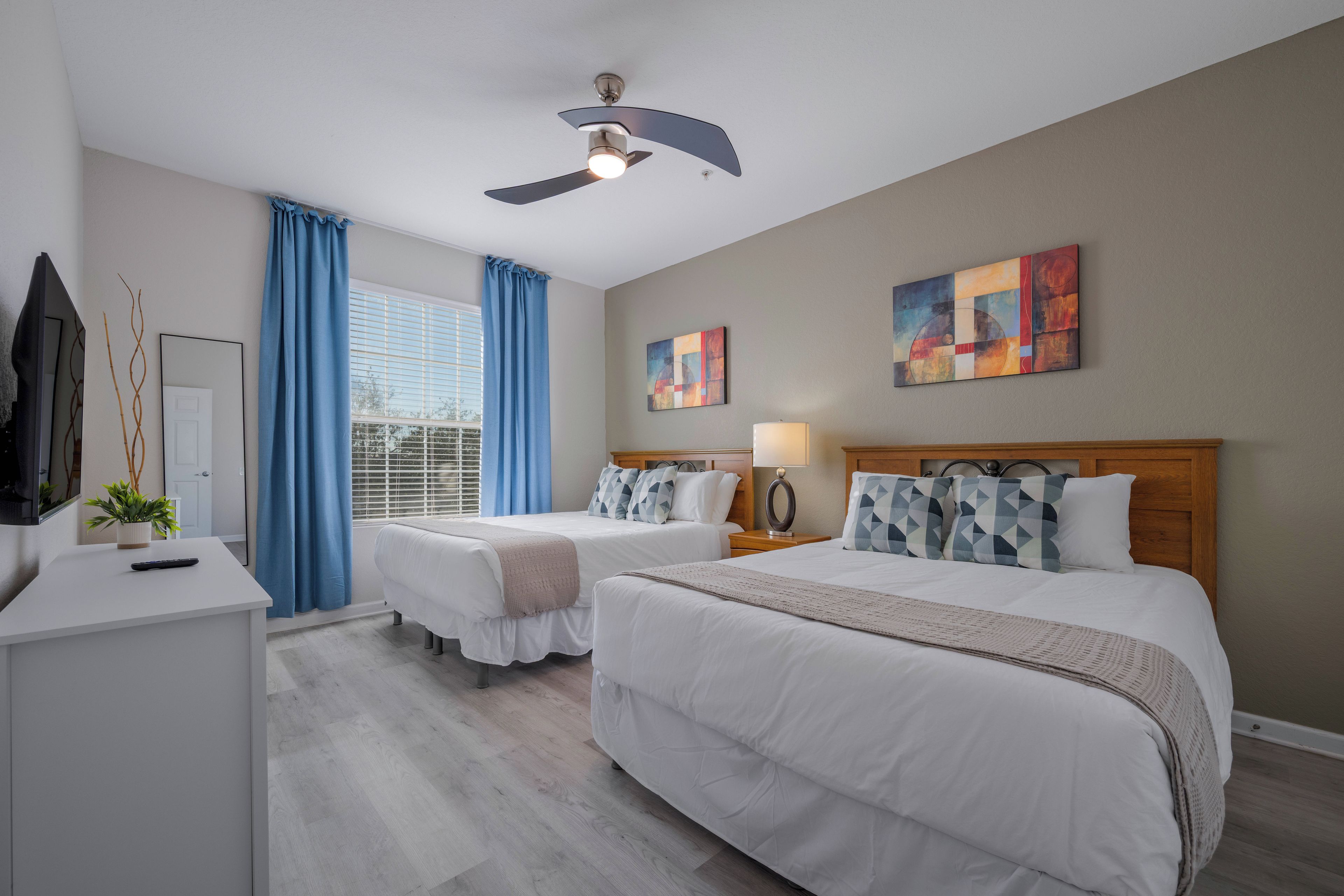 A hotel room with two beds, a desk, a ceiling fan, and artwork on the walls.