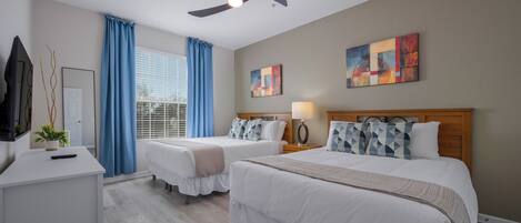 A hotel room with two beds, a desk, a ceiling fan, and artwork on the walls.