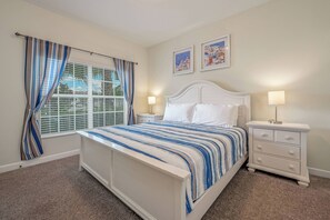 Een slaapkamer met een wit bed, gestreepte gordijnen, een nachtkastje met een lamp en twee ingelijste foto's aan de muur.