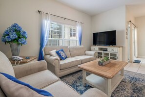 Ein Wohnzimmer mit beiger Couch, einem Holztisch, einem Fernseher auf einer Kommode und einer Vase mit blauen Blumen.
