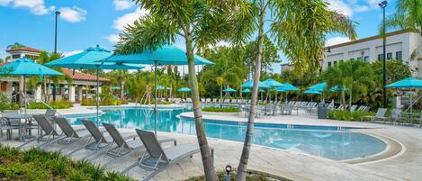 A pool area with lounge chairs and umbrellas, surrounded by palm trees and a building in the background.