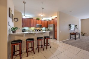 A kitchen with wooden cabinets, a stainless steel refrigerator, a microwave, and a sink. There are three stools and a small table with a plant. - Vc10-208 - 3BR Large Condo Near the Attractions (Orlando)