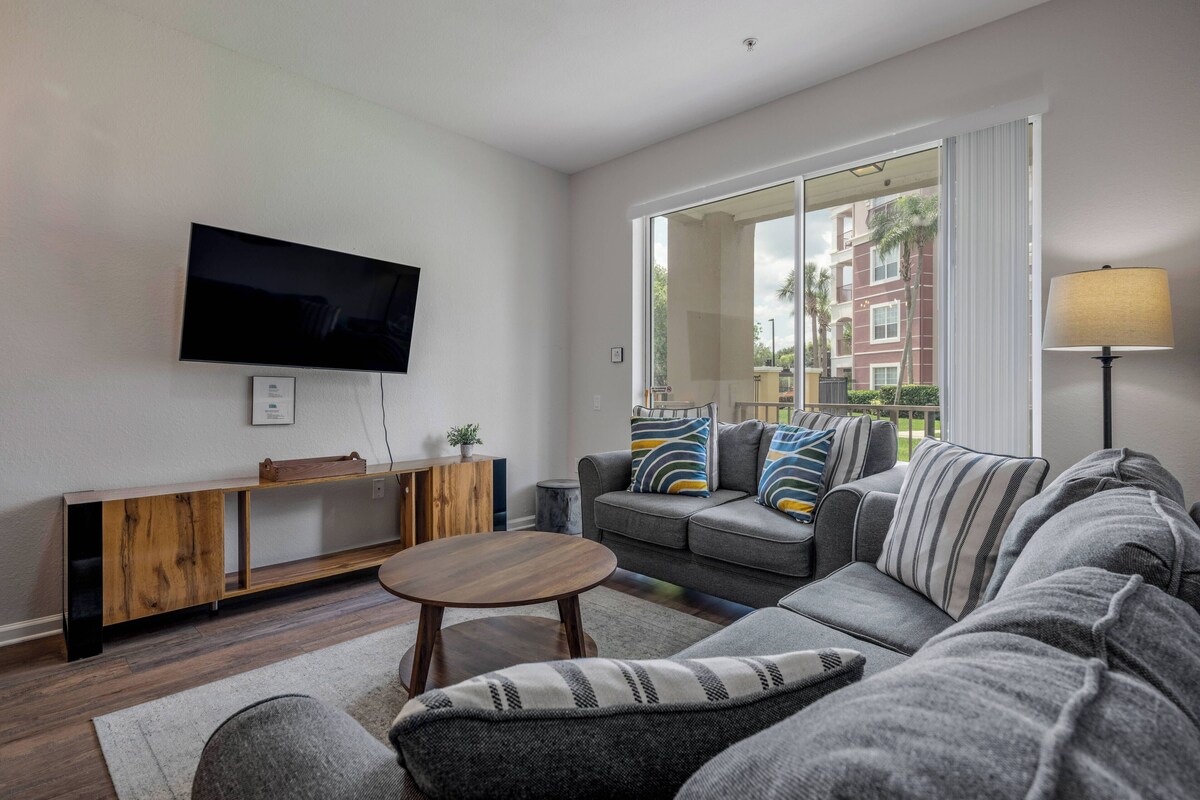 A modern living room with a flat-screen TV, a wooden console table, a grey sofa with striped cushions, and a large window offering a view of buildings and greenery.
