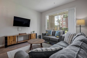 A modern living room with a flat-screen TV, a wooden console table, a grey sofa with striped cushions, and a large window offering a view of buildings and greenery. - Vc8-410 - 3BR Condo With hot tub Near Disney (Orlando)