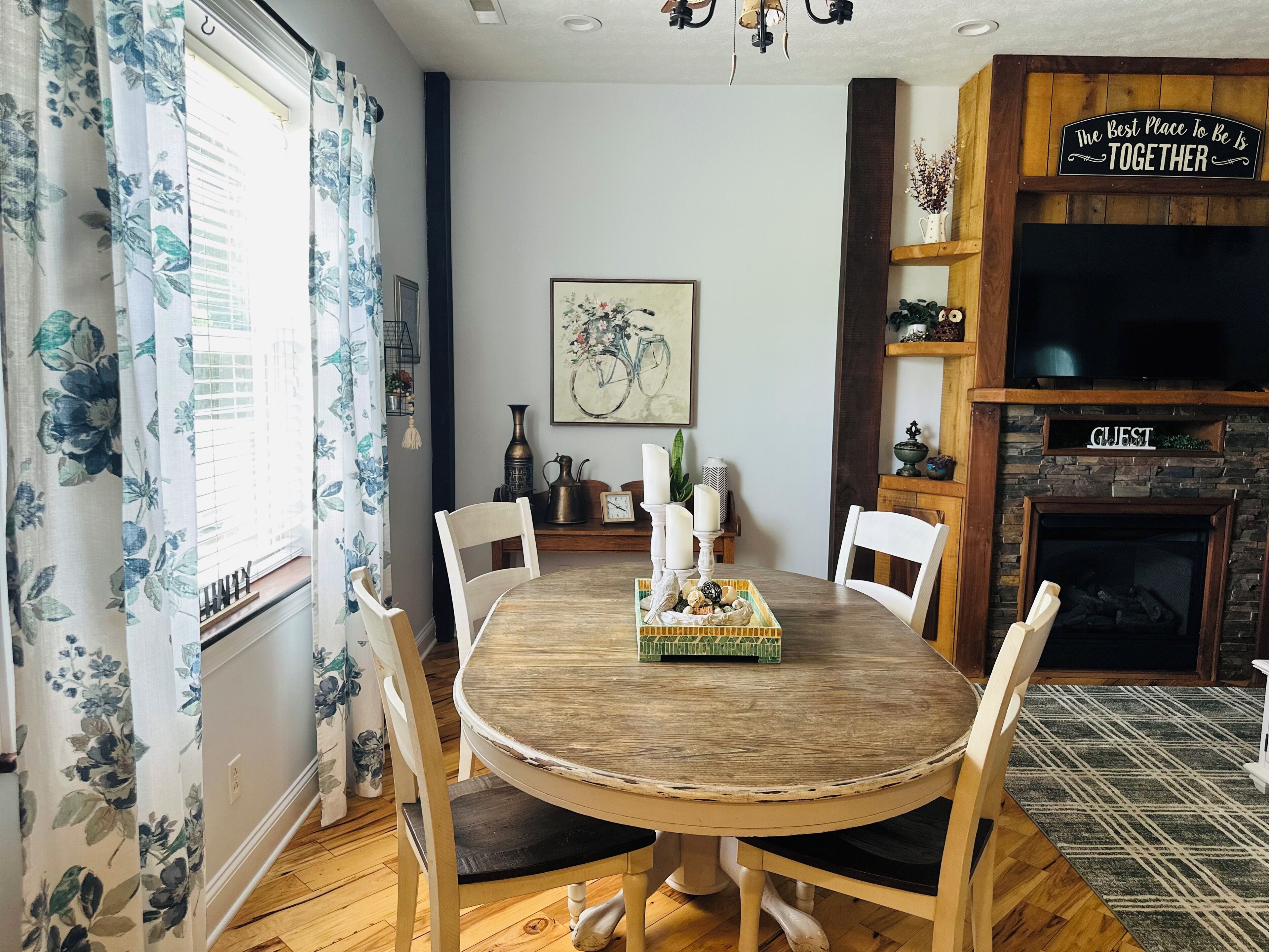 A dining area with a round wooden table, white chairs, a floral curtain, a fireplace, and a 'The Best Place To Be Is Together' sign.