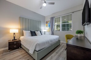 Quarto com cama, mesa de cabeceira, luminária, cadeira, escrivaninha, televisão, janela com persianas e ventilador de teto.