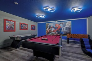 Ein Billardtisch mit roter Spielbahn, ein Tischfußballspiel und zwei gerahmte Poster an der Wand.