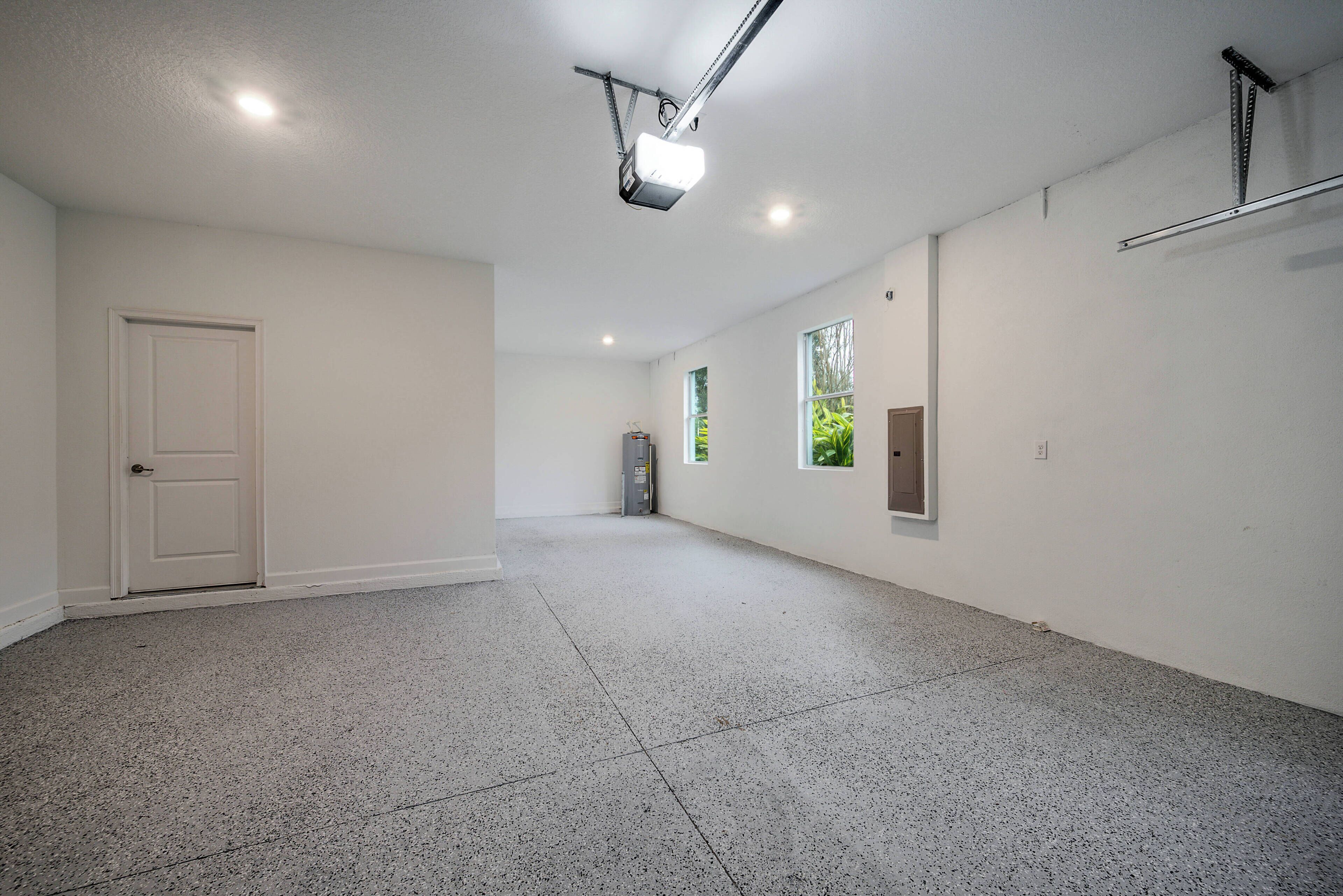 A spacious, well-lit hallway with a door on the left, a projector mounted on the ceiling, and a window with a view of greenery on the right.