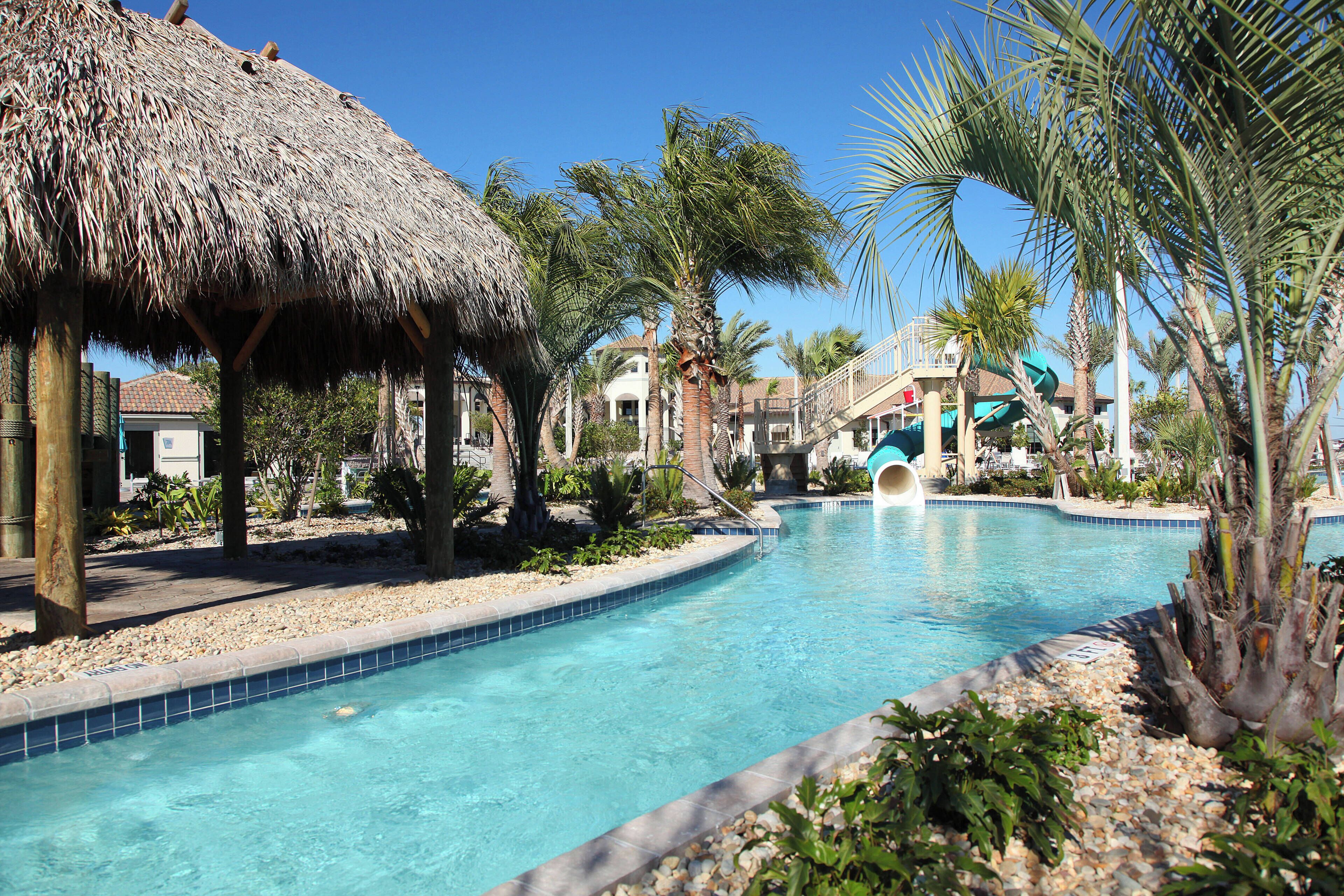 A swimming pool with a thatched roof structure, palm trees, and a water slide.