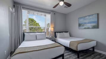 Un dormitorio con dos camas, un ventilador de techo, una ventana con cortinas, una mesita de noche y un cuadro en la pared.