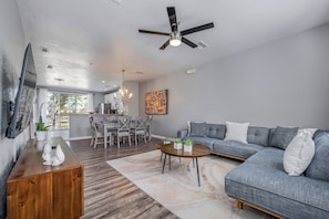 A modern living room with a grey sofa, a wooden coffee table, and a dining area with a chandelier. - Th20 - 3BR House With Resort Access Near Parks (Orlando)