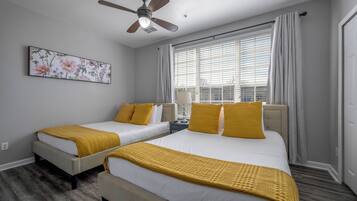 Quarto de hotel com duas camas, ventilador no teto, janela com cortinas e uma obra de arte floral na parede.