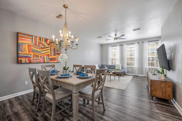 A dining area with a wooden table, chairs, and a chandelier. There is a painting on the wall and a TV mounted on the wall. - Th20 - 3BR House With Resort Access Near Parks (Orlando)