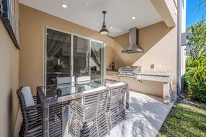 Una terrazza esterna con tavolo in vetro e sedie in vimini, un barbecue e una porta scorrevole che dà su un'area coperta.