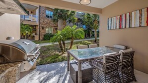An outdoor patio with a grill, wicker furniture, and a view of a residential area with palm trees and a building. - Mvy7632 - 4BR House Near Disney (Kissimmee)