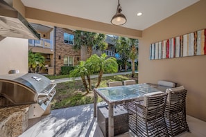 Un patio exterior con una parrilla, mobiliario de mimbre y vistas a una zona residencial con palmeras y un edificio.