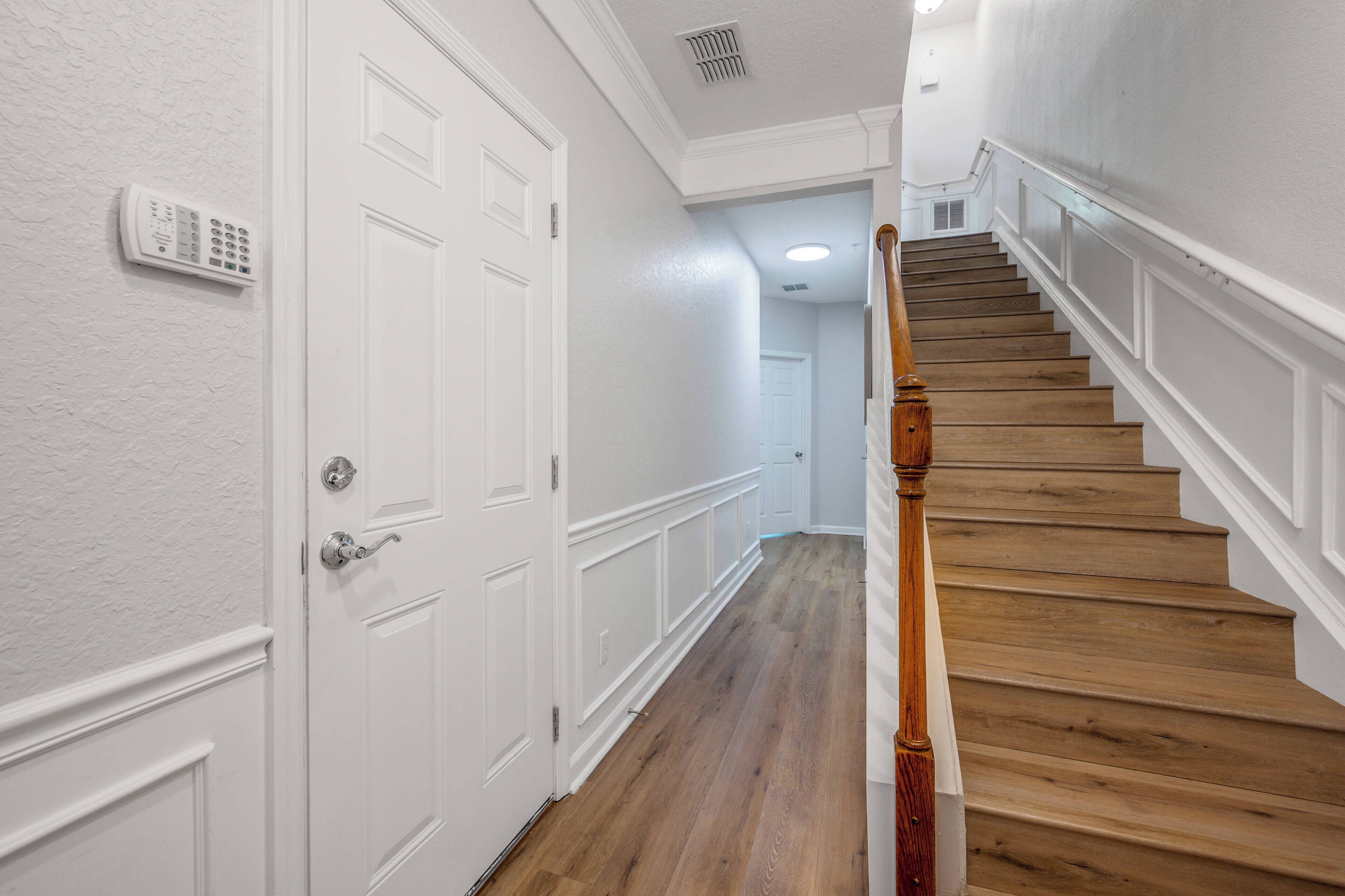 A hallway with a wooden staircase, white walls, and a door with a security keypad.