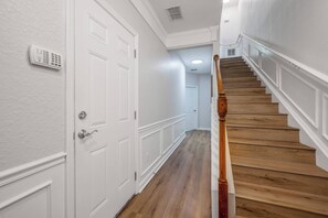 Um corredor com escada de madeira, paredes brancas e porta com teclado de segurança.