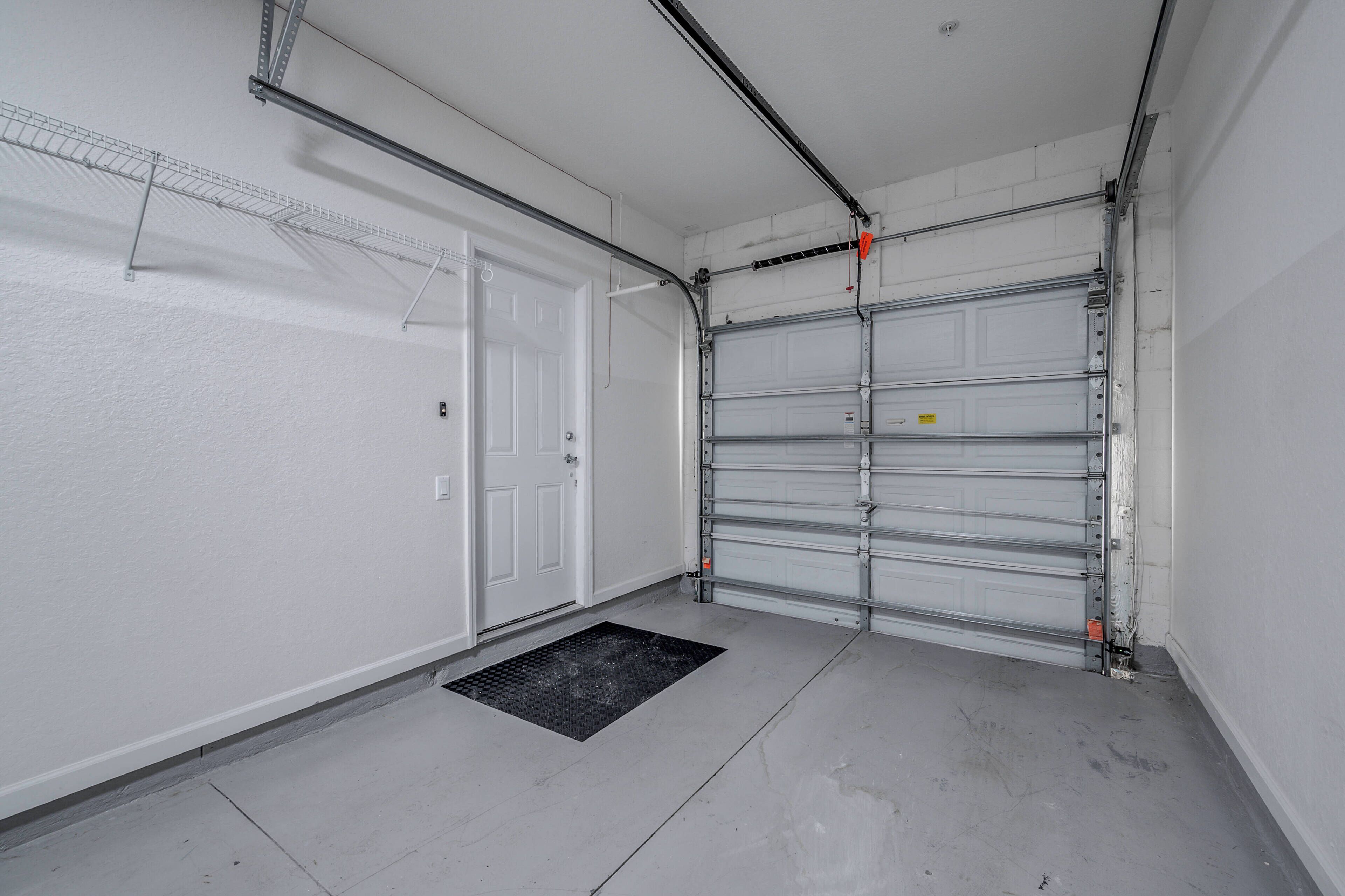 A clean, empty garage with a closed white garage door, a metal shelving unit, and a black mat at the entrance.