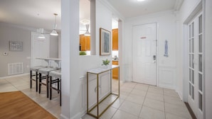 A modern kitchen with white cabinetry, a marble countertop, and a built-in oven. There is a dining area with a bar stool and a small table. - Vc6-105 - 3BR Condo Resort Access Near Disney (Orlando)