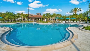 A large outdoor swimming pool with a curved design, surrounded by lounge chairs and umbrellas, and a building with a red-tiled roof in the background. - Vc6-105 - 3BR Condo Resort Access Near Disney (Orlando)