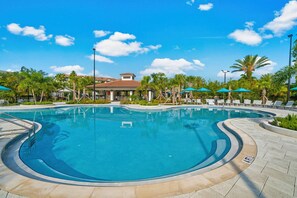 A large outdoor swimming pool with a curved design, surrounded by lounge chairs and umbrellas, and a building with a red-tiled roof in the background. - Vc6-105 - 3BR Condo Resort Access Near Disney (Orlando)