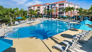 Hotel met een groot zwembad, omringd door ligbedden en parasols, palmbomen en een helderblauwe lucht.