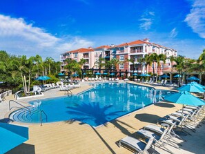 Ein Hotel mit einem großen Pool, umgeben von Liegestühlen und Sonnenschirmen, Palmen und blauem Himmel.