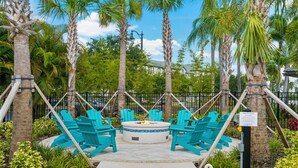 A fire pit area with turquoise Adirondack chairs and a circular seating arrangement surrounded by palm trees and a fence. - Vc12-206 - 3BR Condo Lake View Near Disney (Orlando)