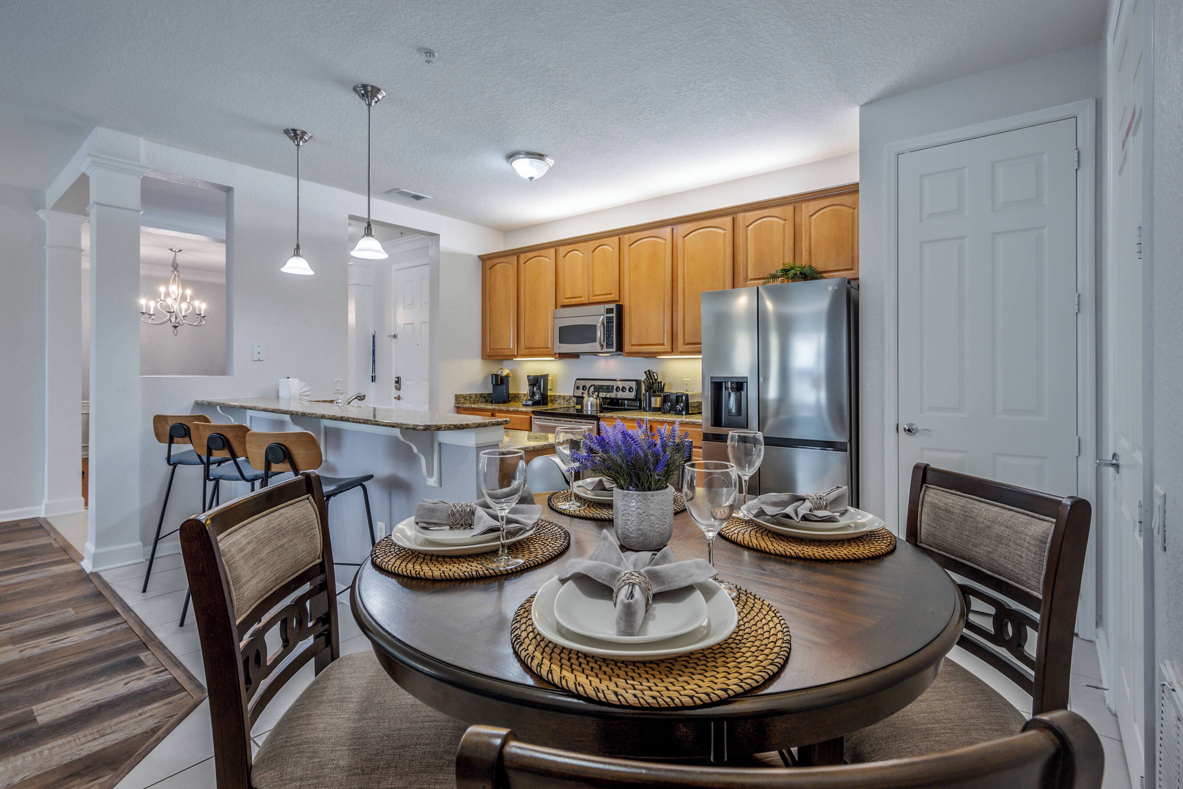 A dining area with a round table set for four, surrounded by wooden chairs. There is a kitchen in the background with wooden cabinets and stainless steel appliances.