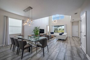 A spacious living area with a glass dining table, comfortable seating, a large window, and a view of the outdoors. - Sl4807 - 5BR House Near Disney Parks Modern Home (Kissimmee)