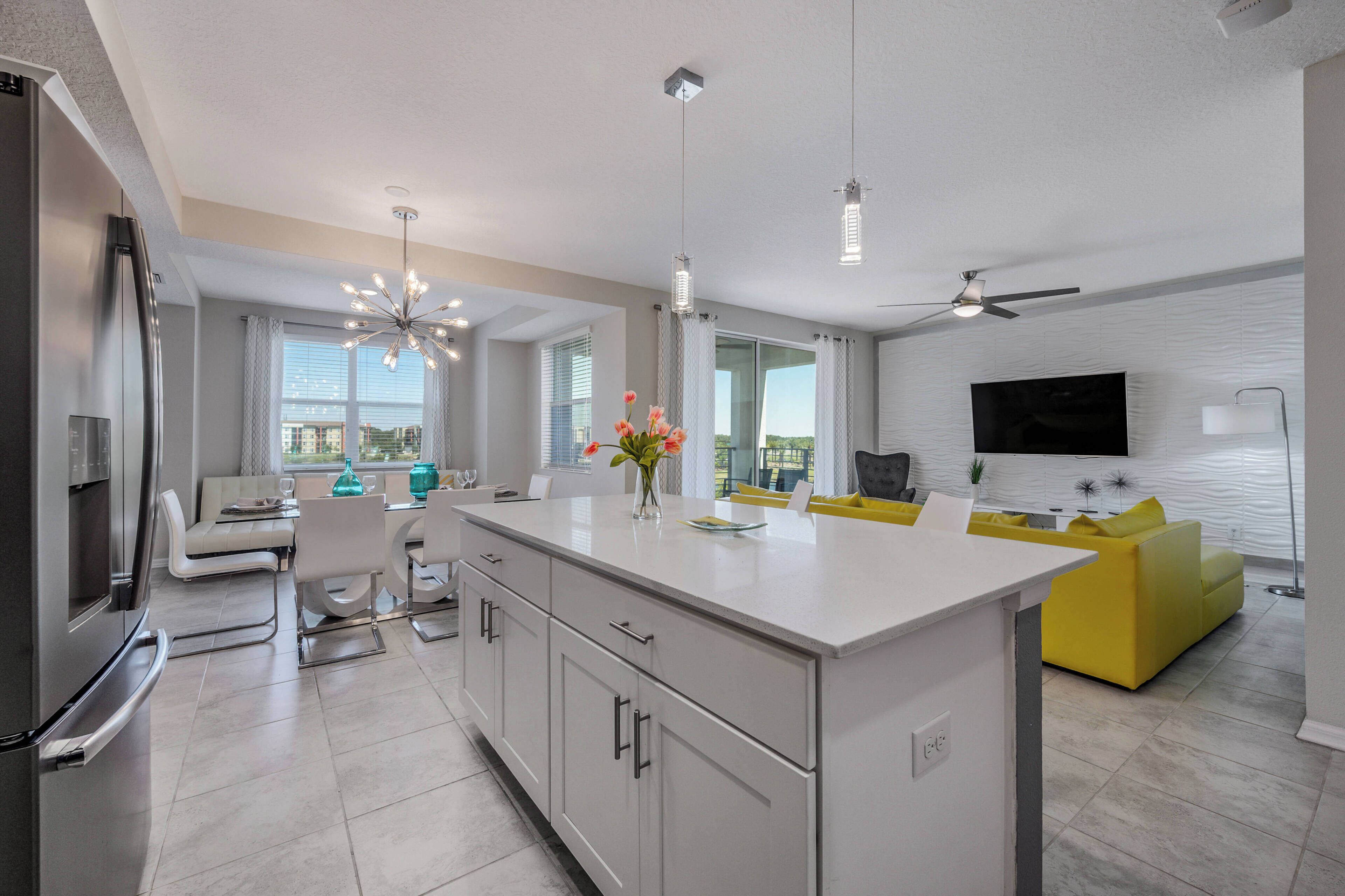 A modern kitchen with a central island, dining area with a sofa, and a living room with a TV and yellow sofa.